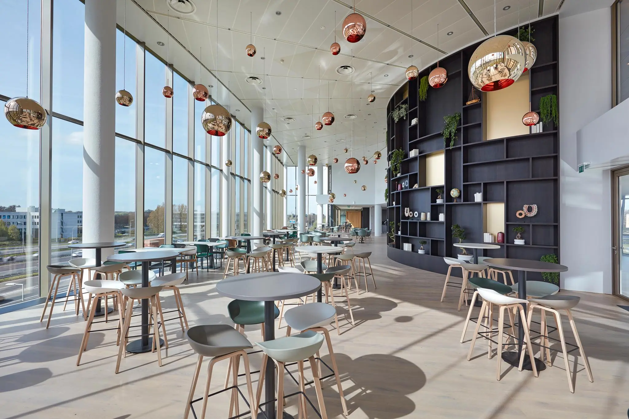 Moderne en open kantoorlounge aan de Fascinatio Boulevard met grote ramen, hoge tafels, designstoelen en een wand met decoratieve objecten en planten.